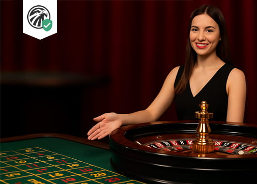 Smiling female live roulette dealer in a black dress gestures toward a roulette wheel and betting table.